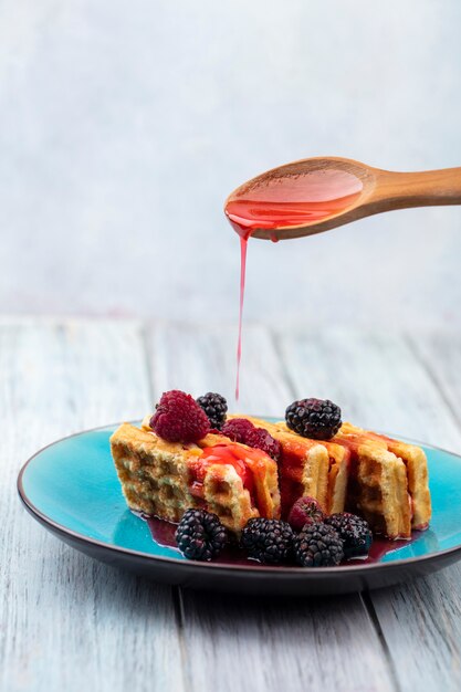 Vorderansicht der süßen Waffeln mit Brombeere auf einem blauen Teller mit einem Holzlöffel mit Sirup auf einer grauen Oberfläche