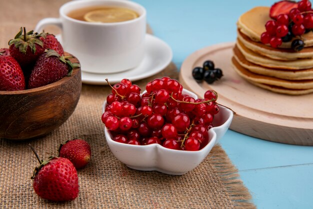 Vorderansicht der Tasse Tee mit einer Zitronenscheibe und Pfannkuchen mit roten und schwarzen Johannisbeeren und Erdbeeren auf einer blauen Oberfläche