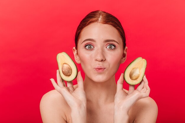 Vorderansicht der verblüfften Ingwerfrau mit Avocado. Studioaufnahme des raffinierten Mädchens lokalisiert auf rotem Hintergrund.