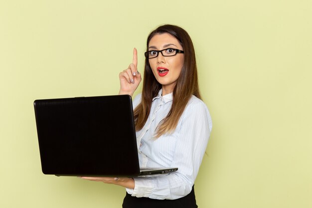 Vorderansicht der weiblichen Büroangestellten im weißen Hemd und im schwarzen Rock, die ihren Laptop auf grüner Wand halten