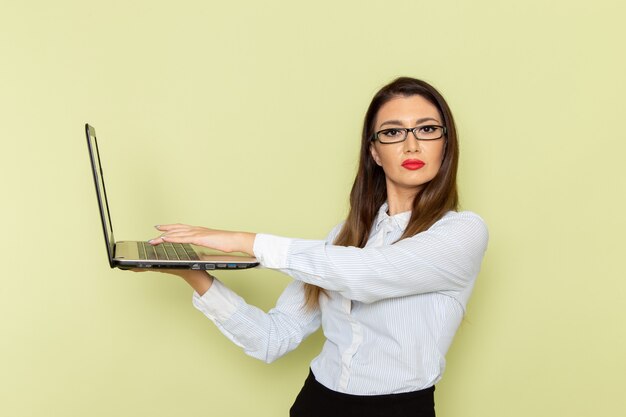 Vorderansicht der weiblichen Büroangestellten im weißen Hemd und im schwarzen Rock unter Verwendung ihres Laptops auf der hellgrünen Wand