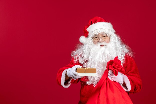 Vorderansicht der Weihnachtsmann, der ein Geschenk aus einer Tasche voller Geschenke für Kinder auf dem roten Gefühl des neuen Jahres hält