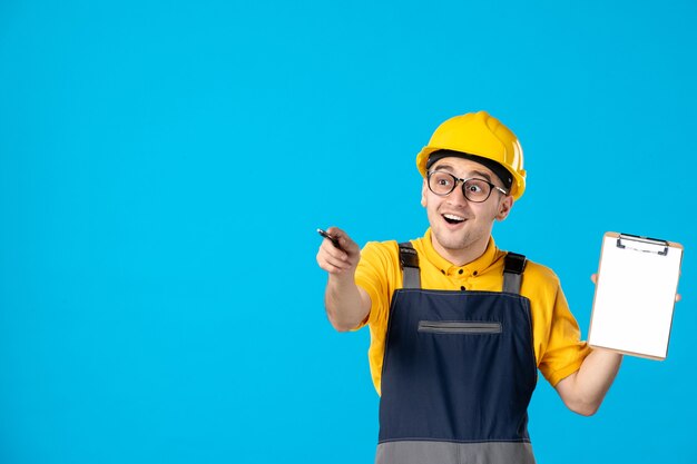 Vorderansicht des aufgeregten männlichen Baumeisters in der Uniform und im Helm mit Notizblock auf einer blauen Wand