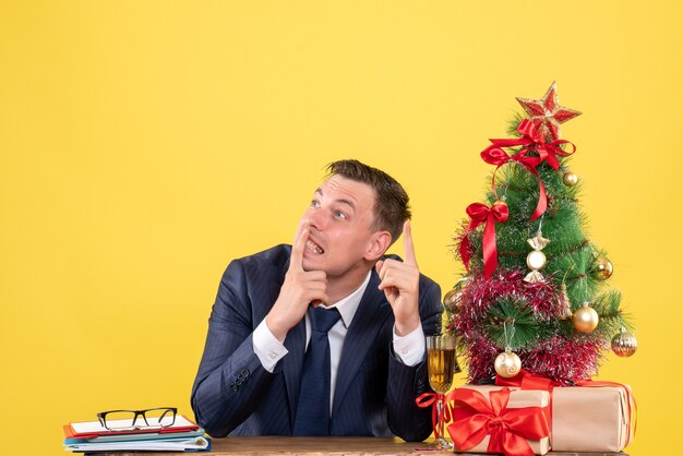 Vorderansicht des denkenden Mannes, der am Tisch nahe Weihnachtsbaum sitzt und auf Gelb präsentiert