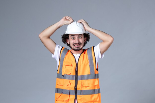 Vorderansicht des denkenden verwirrten männlichen Baumeisters, der Warnweste mit Schutzhelm auf grauer Wellenwand trägt