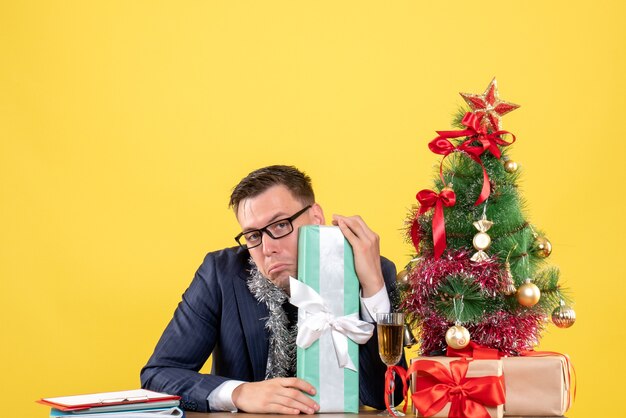 Vorderansicht des depressiven Mannes, der Geschenk hält, das am Tisch nahe Weihnachtsbaum und Geschenken auf Gelb sitzt