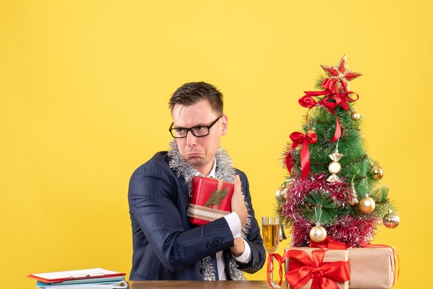 Vorderansicht des depressiven Mannes, der sein Geschenk versteckt, das am Tisch nahe Weihnachtsbaum sitzt und auf Gelb präsentiert