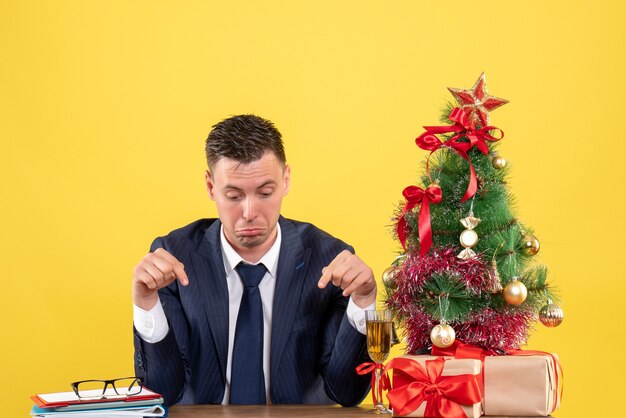 Vorderansicht des depressiven Mannfingers, der den Tisch zeigt, der am Tisch nahe dem Weihnachtsbaum und den Geschenken auf Gelb sitzt