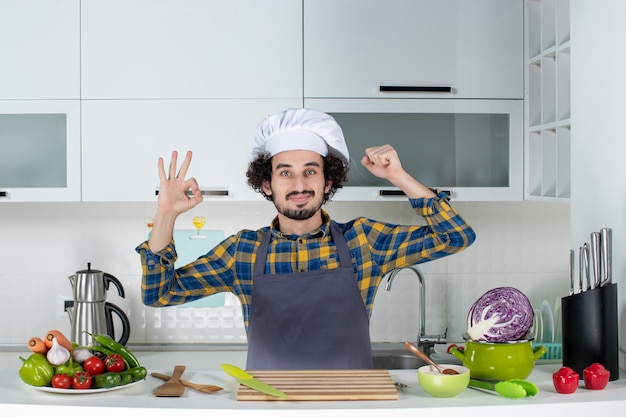 Vorderansicht des ehrgeizigen männlichen Kochs mit frischem Gemüse und Kochen mit Küchengeräten und Brillengeste, die seine Muskeln in der weißen Küche zeigt
