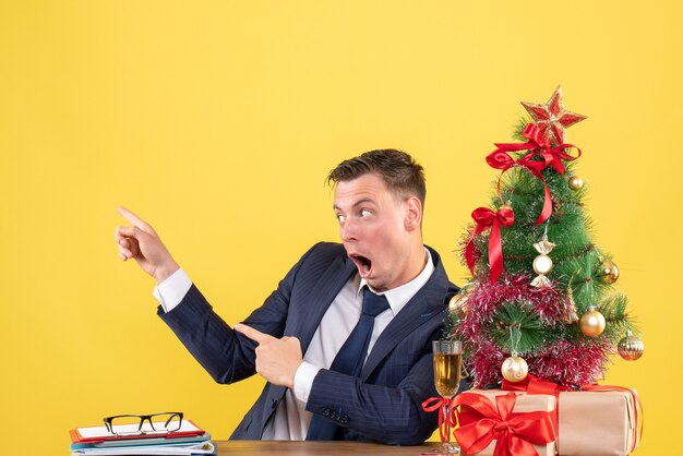 Vorderansicht des erstaunten Mannes, der auf etwas zeigt, das am Tisch nahe Weihnachtsbaum sitzt und auf gelber Wand präsentiert