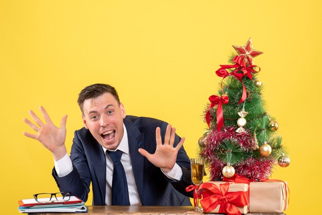 Vorderansicht des fröhlichen Mannes, der Hände öffnet, die am Tisch nahe Weihnachtsbaum sitzen und auf Gelb präsentieren