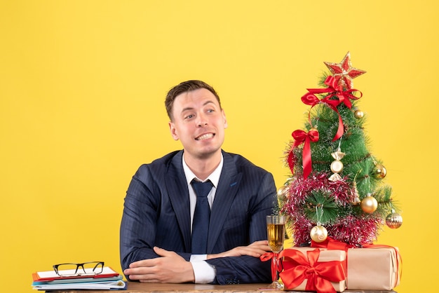 Vorderansicht des gelächelten Mannes, der am Tisch nahe Weihnachtsbaum sitzt und auf Gelb präsentiert