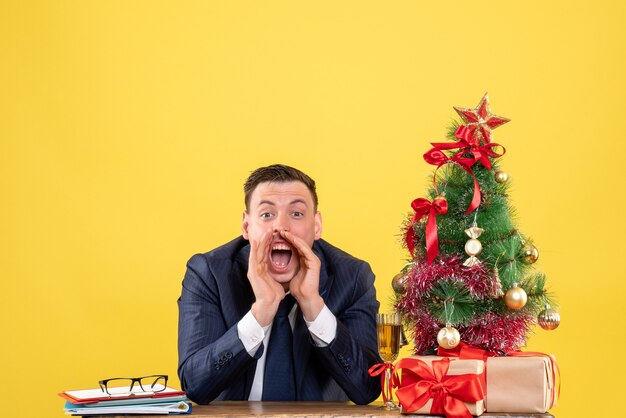 Vorderansicht des gerufenen Mannes, der am Tisch nahe dem Weihnachtsbaum und den Geschenken auf Gelb sitzt.