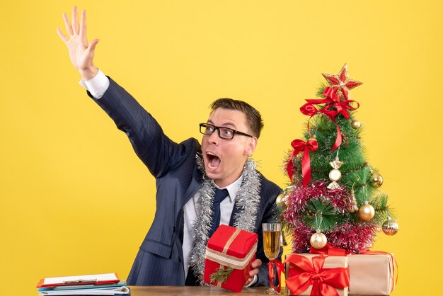 Vorderansicht des gerufenen Mannes, der jemanden anruft, der am Tisch nahe Weihnachtsbaum sitzt und auf Gelb präsentiert.