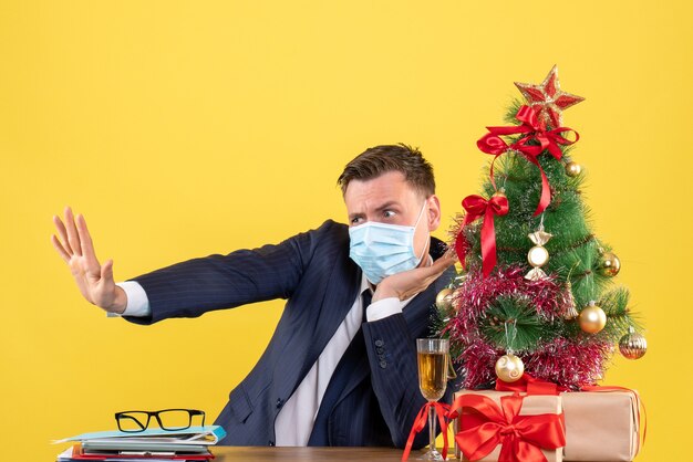 Vorderansicht des Geschäftsmannes, der am Tisch nahe Weihnachtsbaum sitzt und auf Gelb präsentiert.