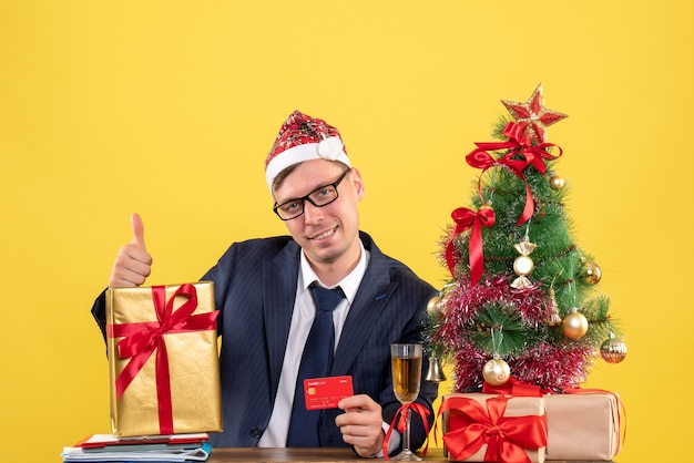 Vorderansicht des Geschäftsmannes, der Daumen hoch Zeichen macht, das am Tisch nahe Weihnachtsbaum und Geschenken auf gelb sitzt