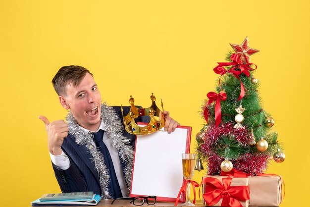 Vorderansicht des Geschäftsmannes, der Daumen hoch Zeichen macht, das am Tisch nahe Weihnachtsbaum und Geschenken auf gelb sitzt