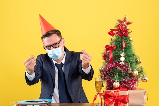 Vorderansicht des Geschäftsmannes, der Geldzeichen mit den Fingern macht, die am Tisch nahe dem Weihnachtsbaum sitzen und auf Gelb präsentieren