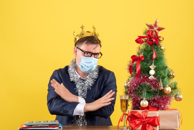 Vorderansicht des Geschäftsmannes, der Hände kreuzt, die am Tisch nahe Weihnachtsbaum sitzen und auf gelber Wand präsentieren