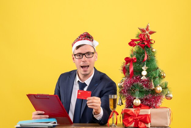 Vorderansicht des Geschäftsmannes, der Karte zeigt, die am Tisch nahe Weihnachtsbaum sitzt und auf Gelb präsentiert