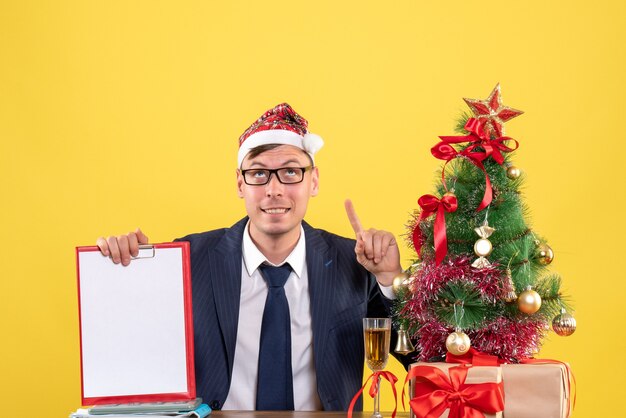 Vorderansicht des Geschäftsmannes, der Klemmbrett hält, das oben sitzt am Tisch nahe Weihnachtsbaum und Geschenke auf Gelb