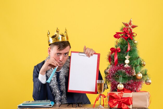 Vorderansicht des Geschäftsmannes, der Krachmacher verwendet, der am Tisch nahe Weihnachtsbaum sitzt und auf Gelb präsentiert