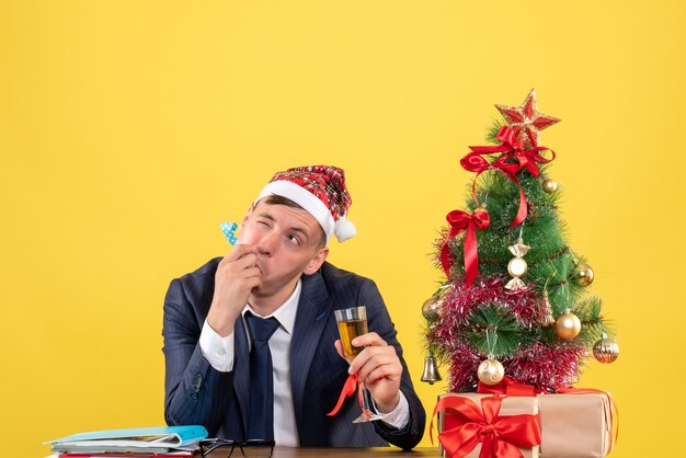 Vorderansicht des Geschäftsmannes, der Krachmacher verwendet, der am Tisch nahe Weihnachtsbaum sitzt und auf Gelb präsentiert