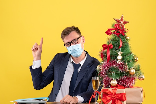 Vorderansicht des Geschäftsmannes, der mit dem Finger zurück zeigt, der am Tisch nahe Weihnachtsbaum sitzt und auf Gelb präsentiert