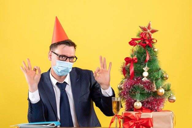 Vorderansicht des Geschäftsmannes, der ok Zeichen macht, das am Tisch nahe Weihnachtsbaum sitzt und auf Gelb präsentiert