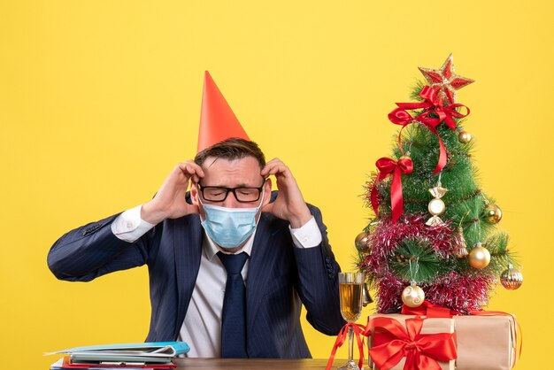 Vorderansicht des Geschäftsmannes, der seinen Kopf hält, der am Tisch nahe Weihnachtsbaum sitzt und auf Gelb präsentiert