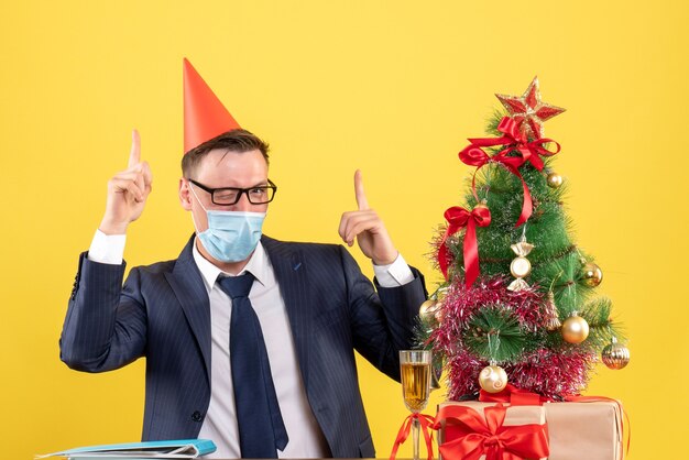 Vorderansicht des Geschäftsmannes mit dem blinkenden Auge der medizinischen Maske, das am Tisch nahe dem Weihnachtsbaum sitzt und auf Gelb präsentiert