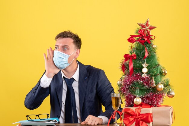 Vorderansicht des Geschäftsmannes mit großen Augen, der am Tisch nahe Weihnachtsbaum sitzt und auf Gelb präsentiert