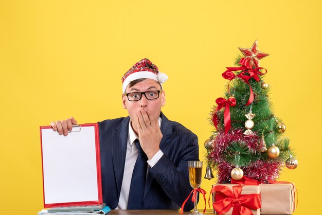 Vorderansicht des Geschäftsmannes mit großen Augen, der Clipoard hält, der am Tisch nahe Weihnachtsbaum sitzt und auf Gelb präsentiert