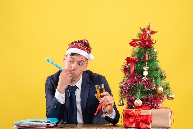 Vorderansicht des Geschäftsmannes mit Krachmacher, der Champagner hält, der am Tisch nahe dem Weihnachtsbaum sitzt und auf Gelb präsentiert