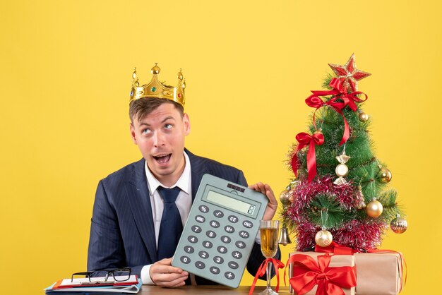 Vorderansicht des Geschäftsmannes mit Kronenhalterechner, der am Tisch nahe Weihnachtsbaum und Geschenken auf Gelb sitzt