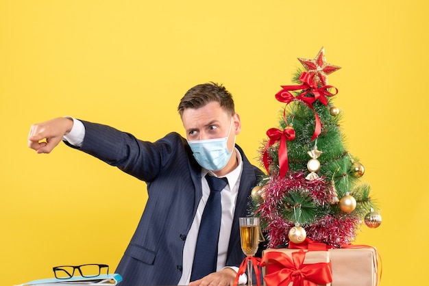 Vorderansicht des Geschäftsmannes mit Maske, die auf jemanden zeigt, der am Tisch nahe Weihnachtsbaum sitzt und auf Gelb präsentiert