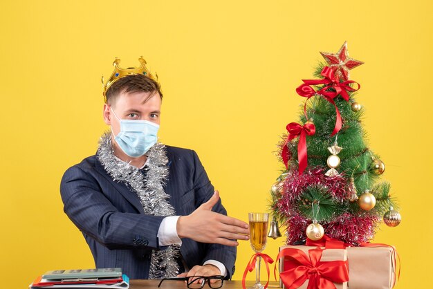 Vorderansicht des Geschäftsmannes mit Maske, die Hand gibt, die am Tisch nahe Weihnachtsbaum sitzt und auf Gelb präsentiert