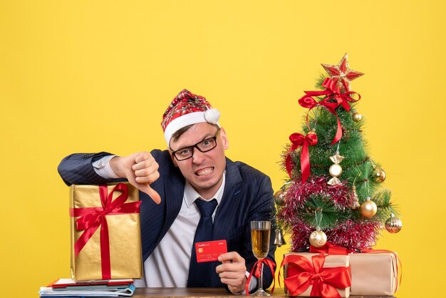 Vorderansicht des Geschäftsmannes mit Weihnachtsmannhut, der Daumen unten Zeichen macht, das am Tisch nahe Weihnachtsbaum sitzt und auf Gelb präsentiert