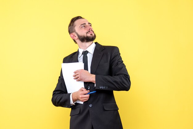 Vorderansicht des Geschäftsmanns des jungen Mannes, der Anzug schaut und über neue Ideen nachdenkt, die leeres Papier mit einem Stift auf Gelb halten