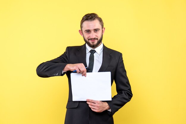 Vorderansicht des Geschäftsmanns des jungen Mannes in einem Anzug, der lächelt und leeres Papier in der Mitte auf gelb hält