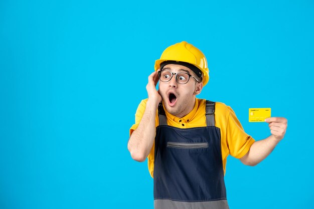 Vorderansicht des geschockten männlichen Baumeisters in der Uniform und im Helm mit Kreditkarte auf einem Blau