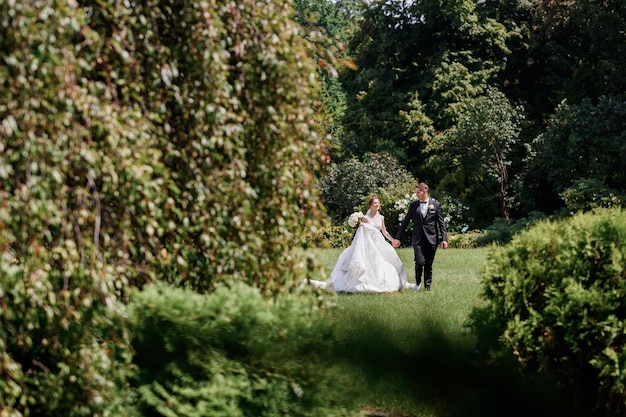 Vorderansicht des glücklichen Ehepaares, das sich an den Händen hält und während des Hochzeitstages in der Sommersaison zusammen durch den wunderschönen Park geht