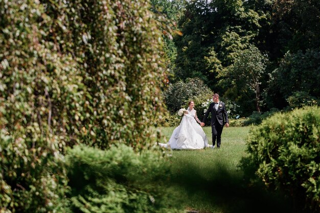 Vorderansicht des glücklichen Ehepaares, das sich an den Händen hält und während des Hochzeitstages in der Sommersaison zusammen durch den wunderschönen Park geht