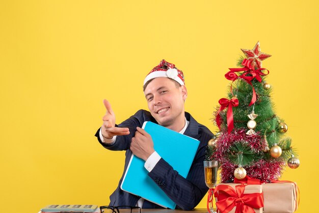 Vorderansicht des glücklichen Geschäftsmannes, der Hand gibt, die am Tisch nahe Weihnachtsbaum sitzt und auf Gelb präsentiert