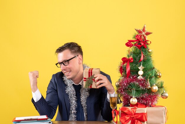 Vorderansicht des glücklichen jungen Mannes, der am Tisch nahe Weihnachtsbaum sitzt und auf Gelb präsentiert