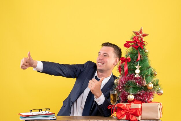 Vorderansicht des glücklichen jungen Mannes, der Daumen oben Zeichen macht, das am Tisch nahe Weihnachtsbaum und Geschenke auf Gelb sitzt