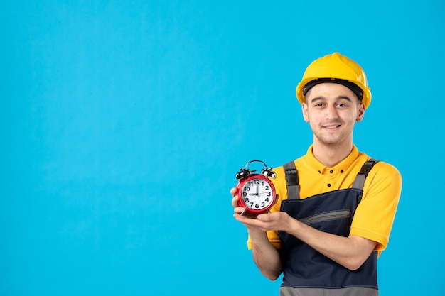 Vorderansicht des glücklichen männlichen Arbeiters in der Uniform mit Uhren auf der blauen Wand