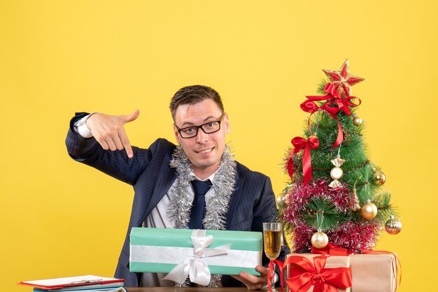 Vorderansicht des glücklichen Mannes, der auf Geschenk zeigt, das am Tisch nahe Weihnachtsbaum sitzt und auf Gelb präsentiert