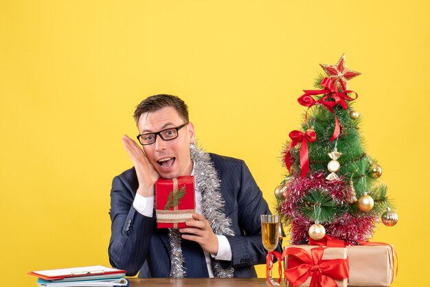 Vorderansicht des glücklichen Mannes, der Geschenk hält, das am Tisch nahe Weihnachtsbaum und Geschenken auf Gelb sitzt