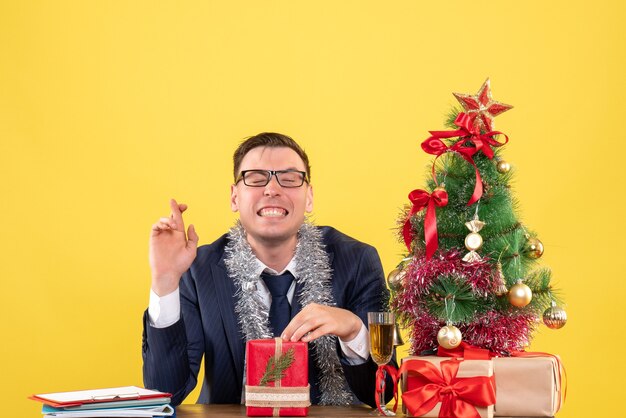 Vorderansicht des glücklichen Mannes, der Glückszeichen macht, das am Tisch nahe Weihnachtsbaum sitzt und auf Gelb präsentiert
